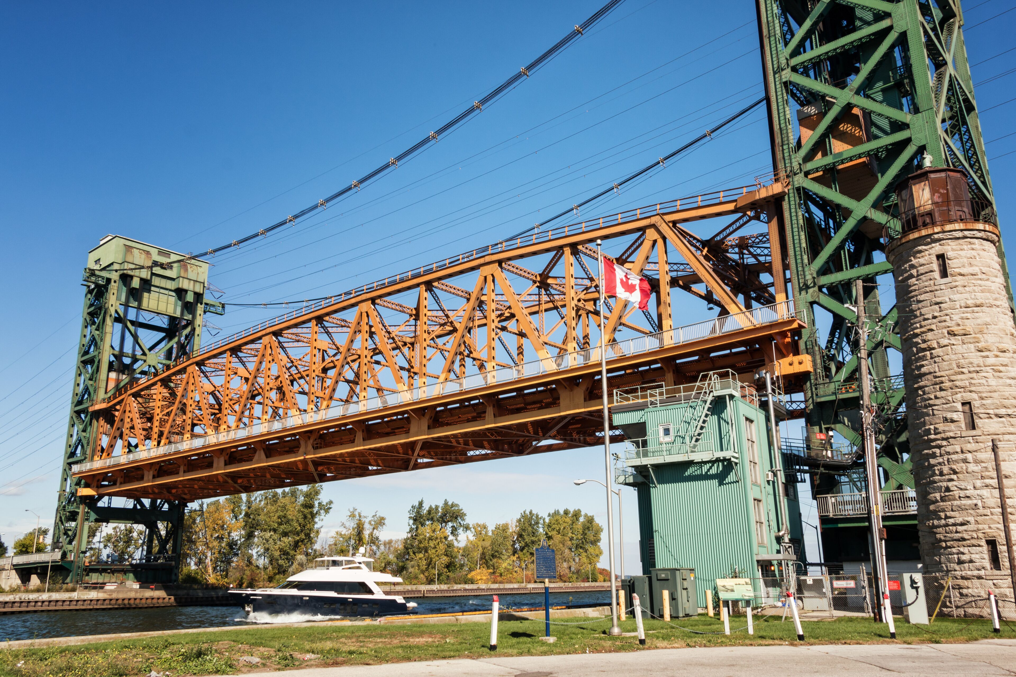 A steel bridge with a Canadian flag, a boat on the water below, and a green industrial structure to the right.
