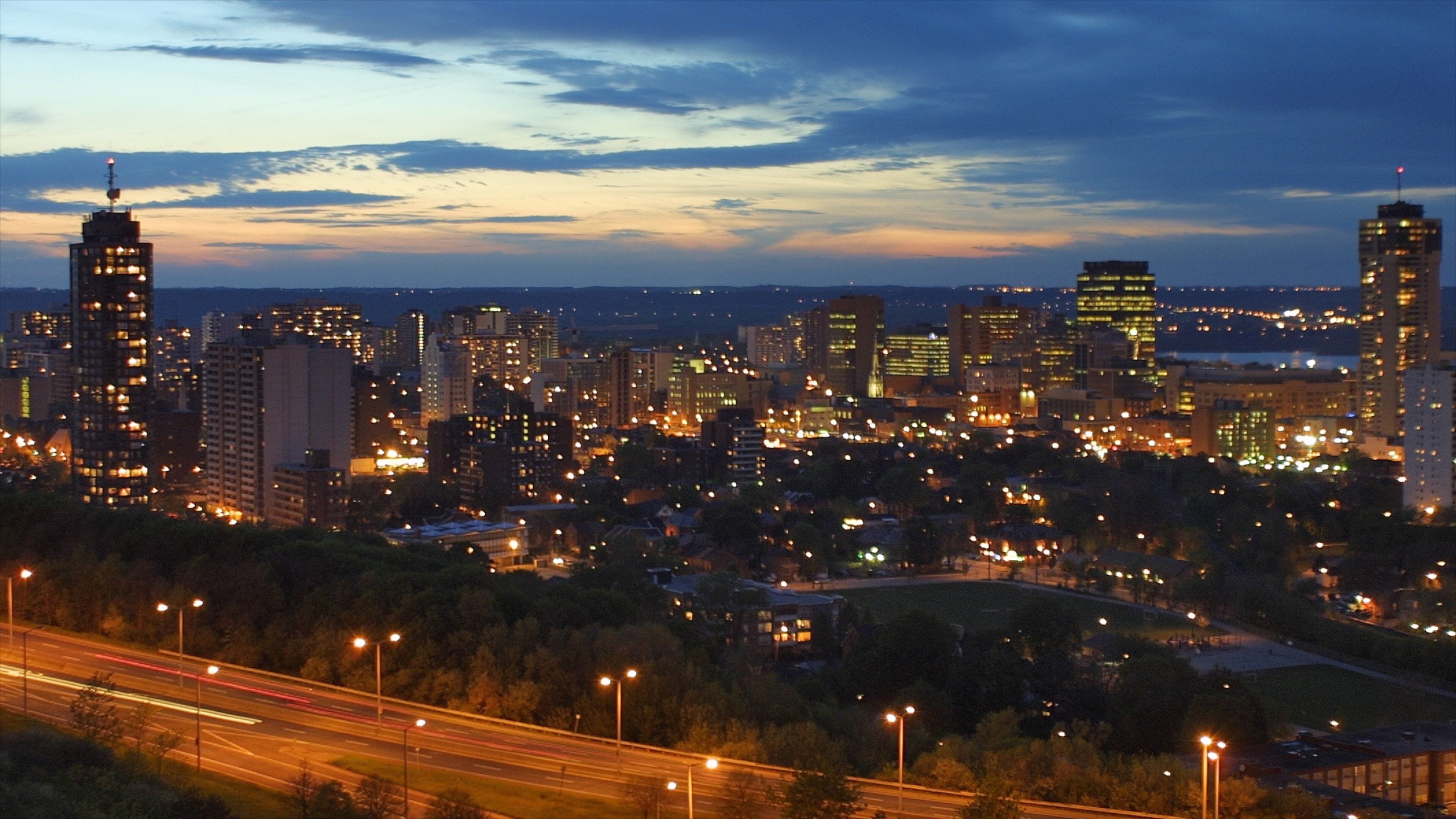 A city skyline at dusk with illuminated buildings and a highway in the foreground.