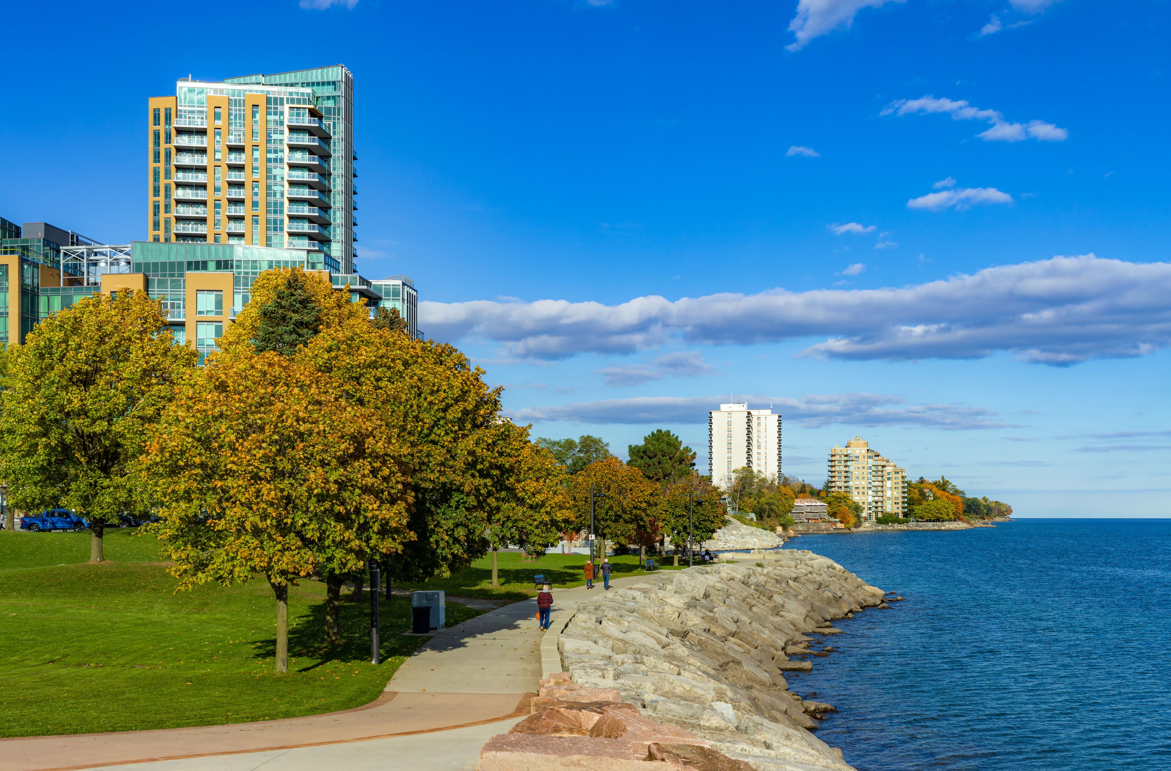 A waterfront promenade with a stone wall, autumn trees, and modern buildings.
