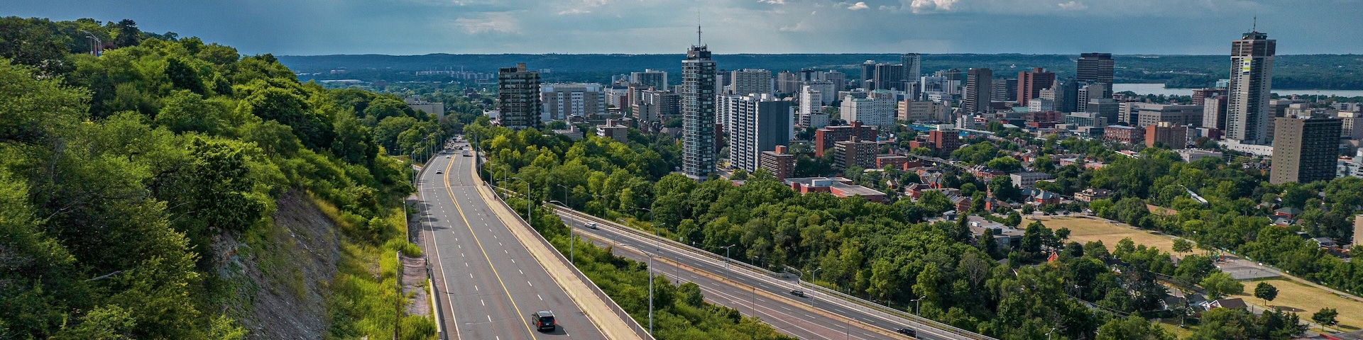 A highway with multiple lanes, surrounded by greenery and urban buildings in the distance.
