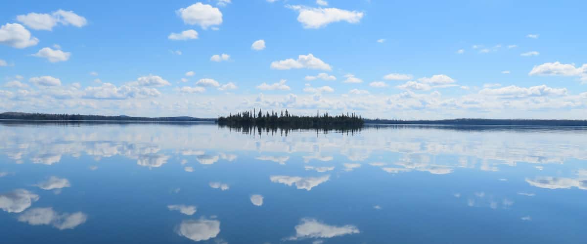 A calm lake with a small island of trees in the center, surrounded by a clear blue sky with scattered clouds.