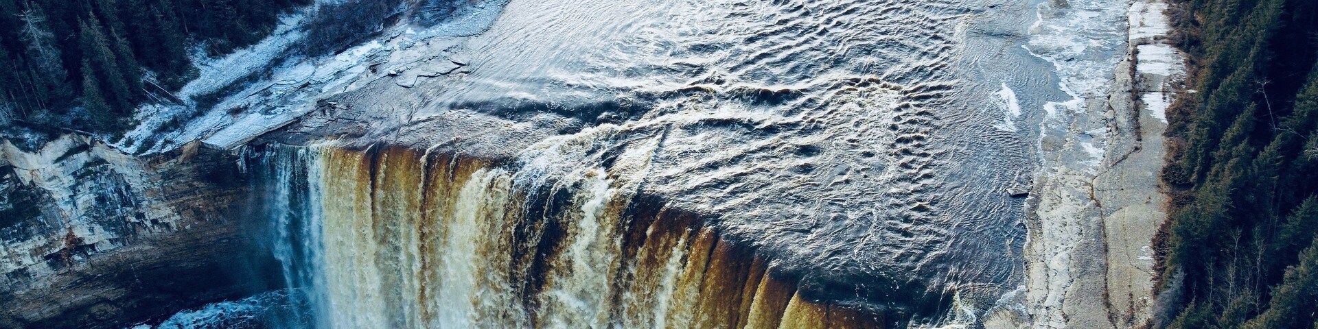 Uma cascata despenca de um penhasco rochoso, cercada por uma floresta densa.
