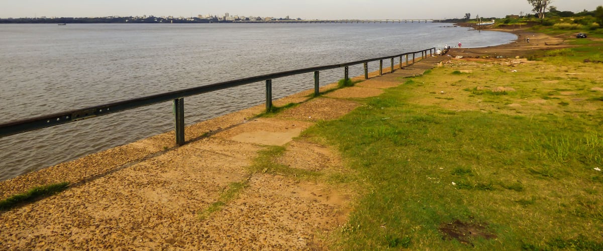 A riverside walkway with a metal railing, grassy banks, and a distant city skyline.