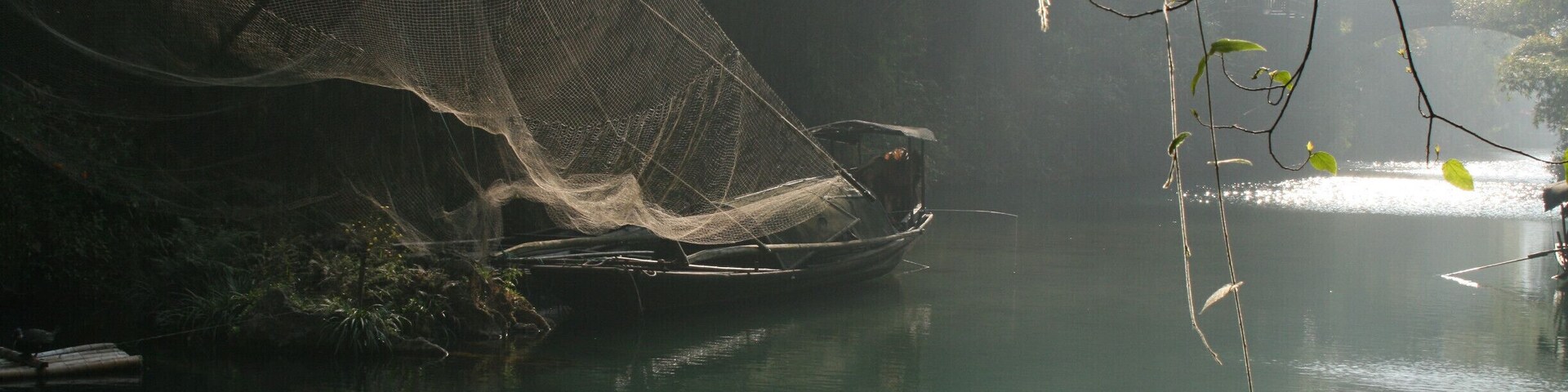 Un bote con una red en un río tranquilo, un puente al fondo y follaje en primer plano.