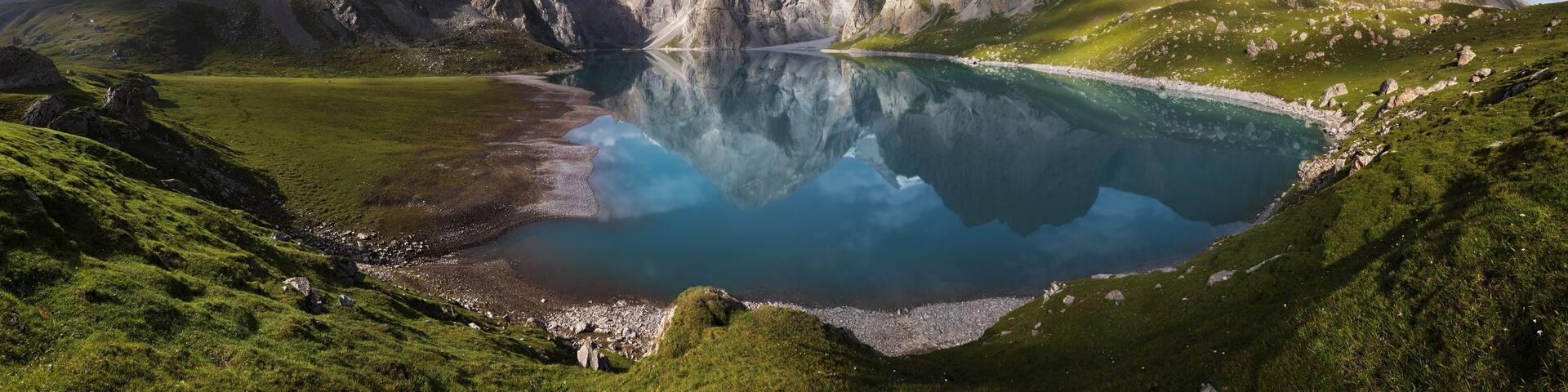 A mountain lake with clear reflections, surrounded by rugged peaks and green hills.