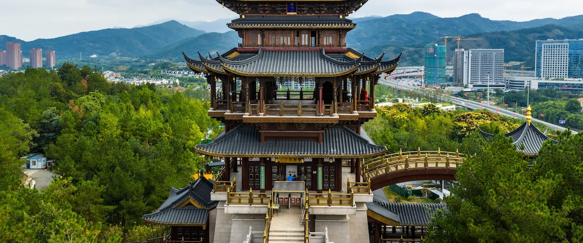 Une pagode traditionnelle à plusieurs niveaux, ornée de motifs dorés, entourée d’une végétation luxuriante et se dressant sur fond de bâtiments modernes et de montagnes.