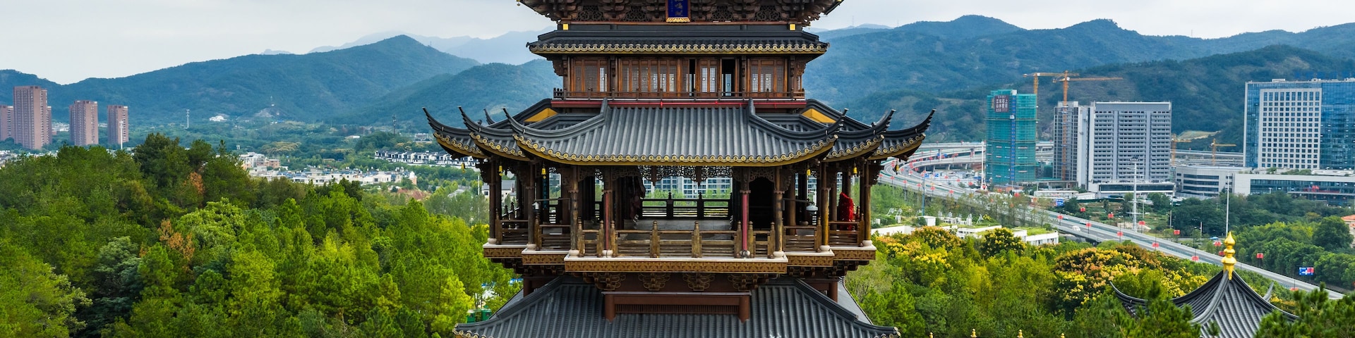 Una pagoda tradicional de varios niveles con adornos dorados, rodeada de exuberante vegetación y con un fondo de edificios modernos y montañas.