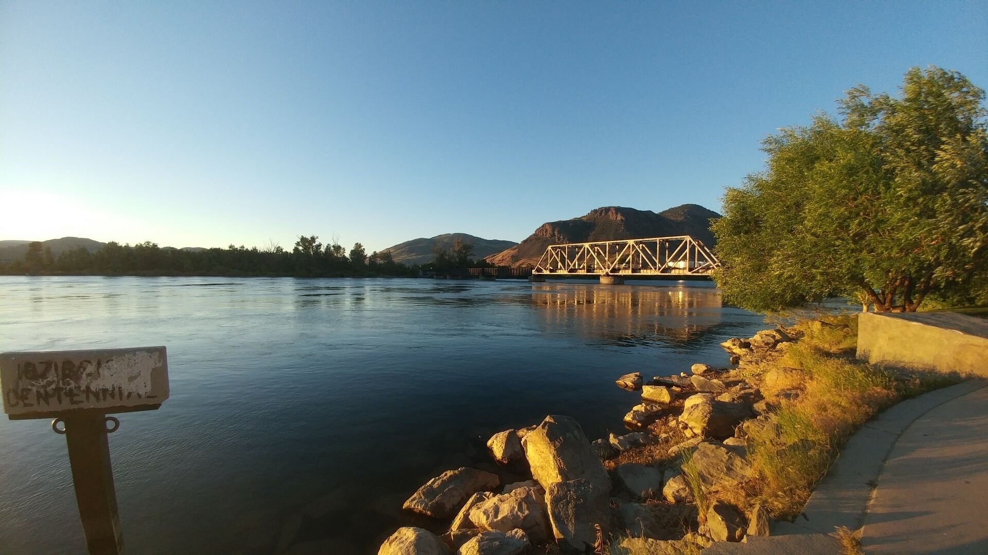 A calm river with a metal bridge, rocky shore, and a signboard.