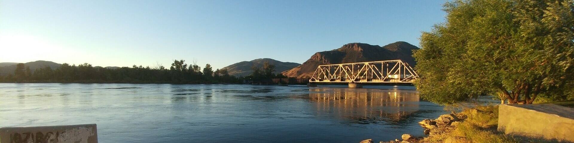 A calm river with a metal bridge, rocky shore, and a signboard.
