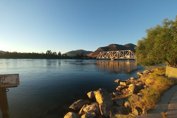 A calm river with a metal bridge, rocky shore, and a signboard.