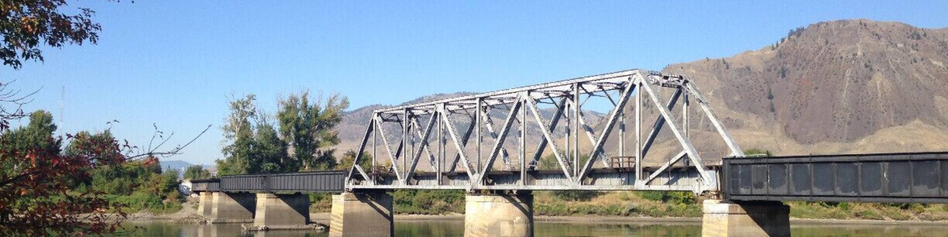A steel truss bridge over a calm river with mountains in the background.