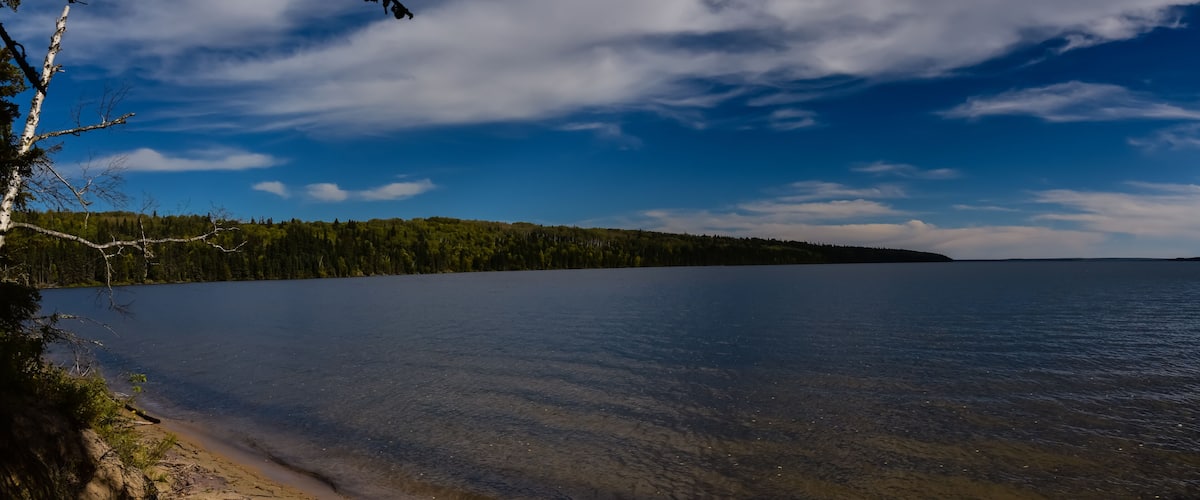 A calm lake with clear water, surrounded by a forested shoreline and a clear blue sky.