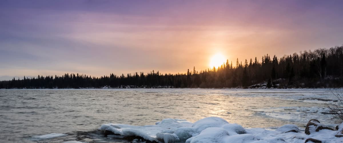 A frozen river with ice formations and a forest in the background during sunset.