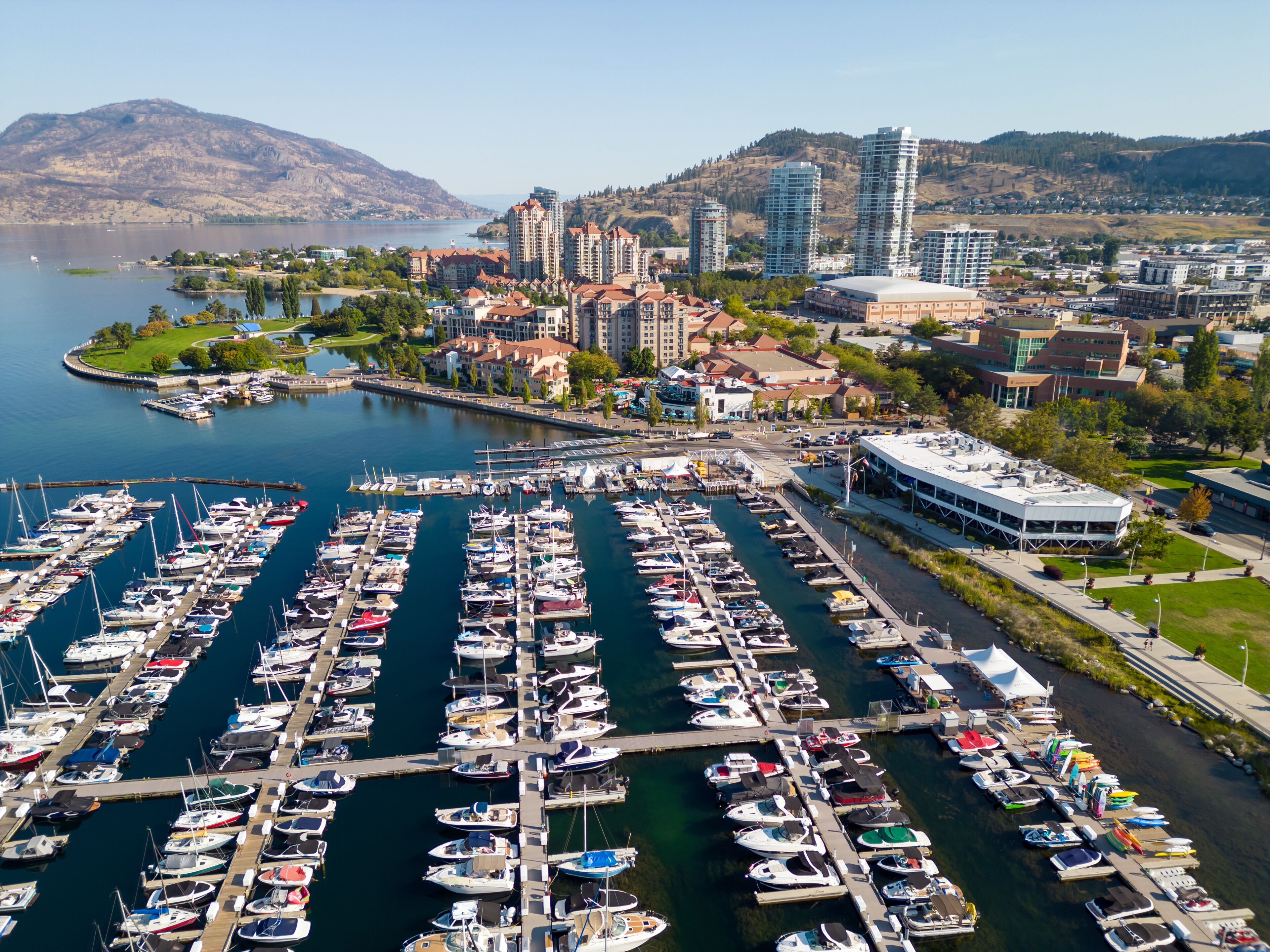 A marina with numerous boats docked, a cityscape with modern buildings, and a mountainous backdrop.