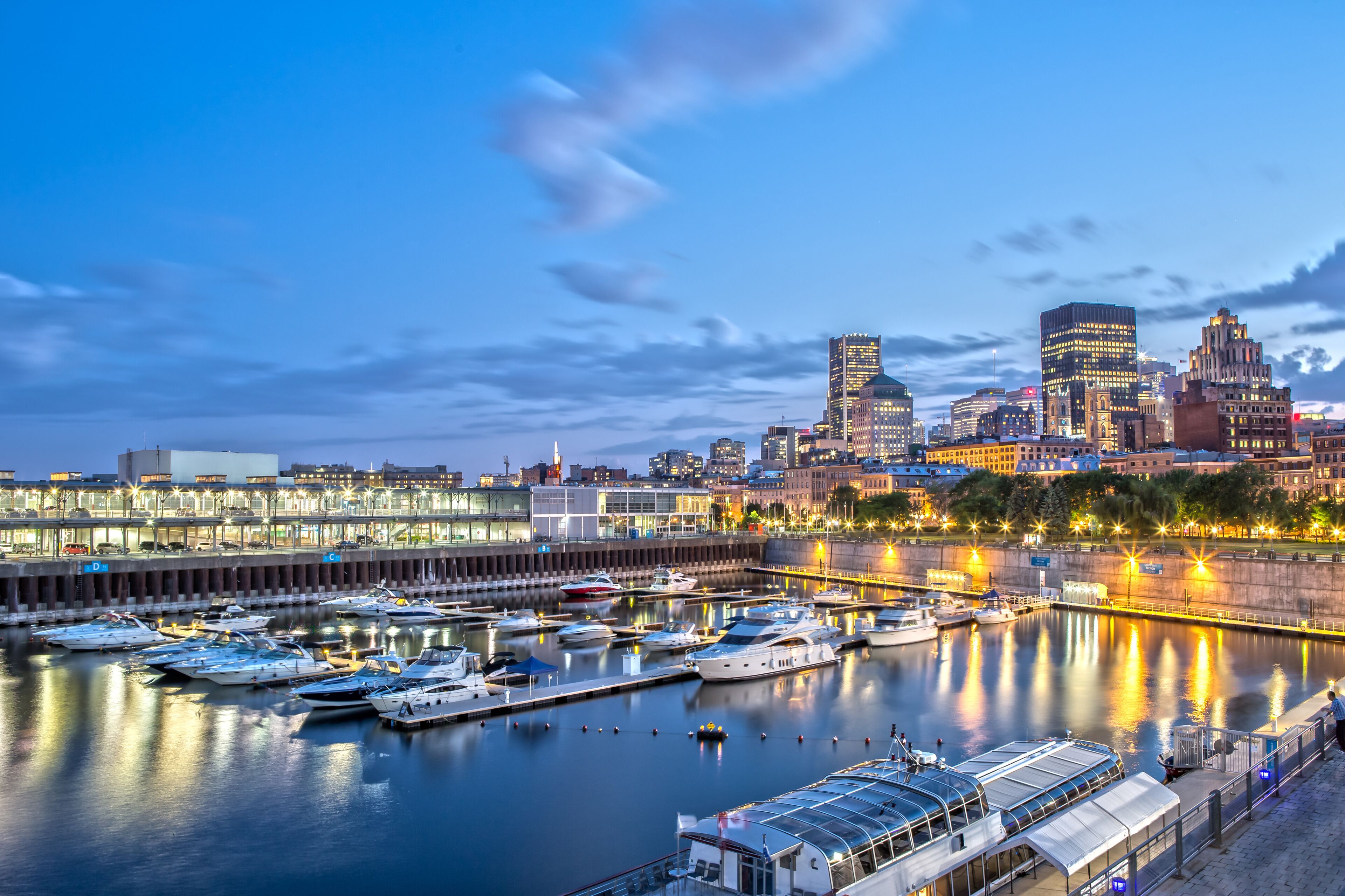 Marina com barcos atracados, horizonte urbano com edifícios iluminados e calçadão à beira-mar.