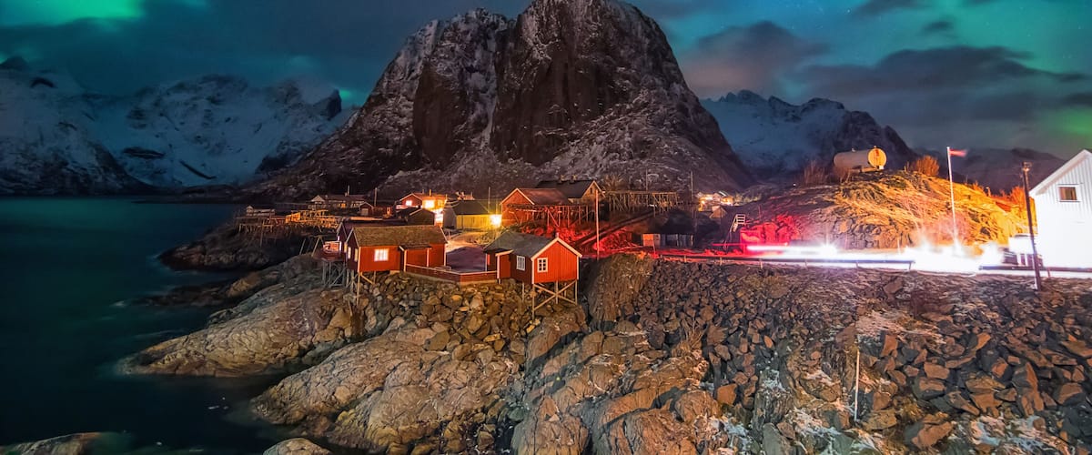 A coastal village with a prominent mountain, illuminated buildings, and a colorful aurora display.