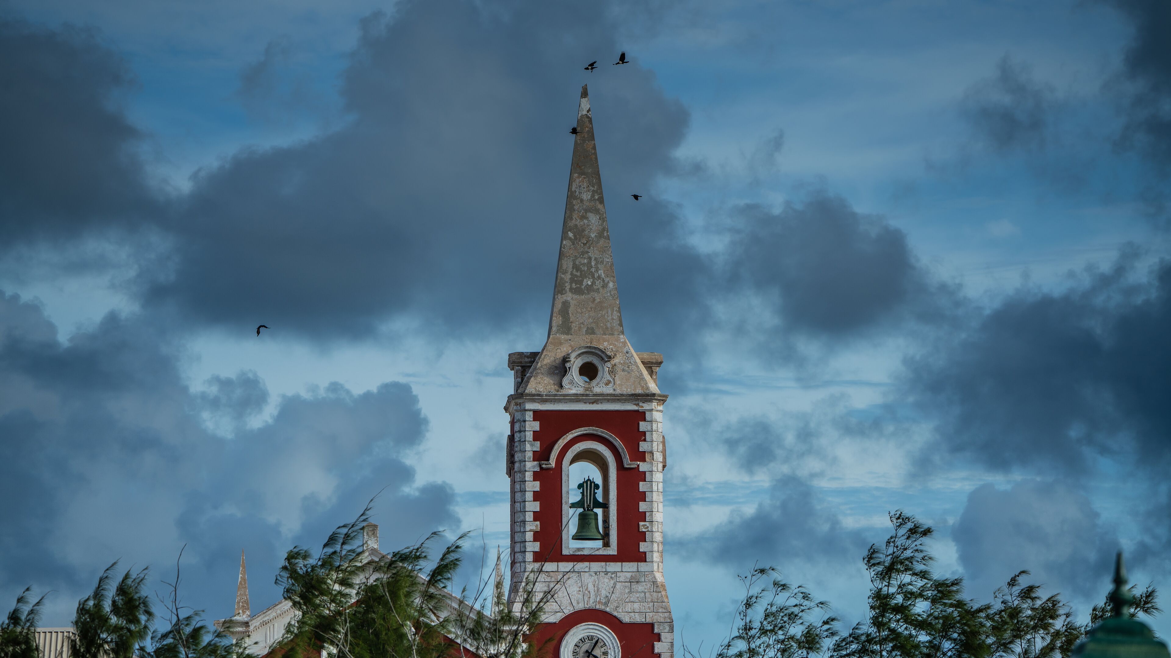 A church tower with a clock and a bell, surrounded by trees and a cloudy sky.