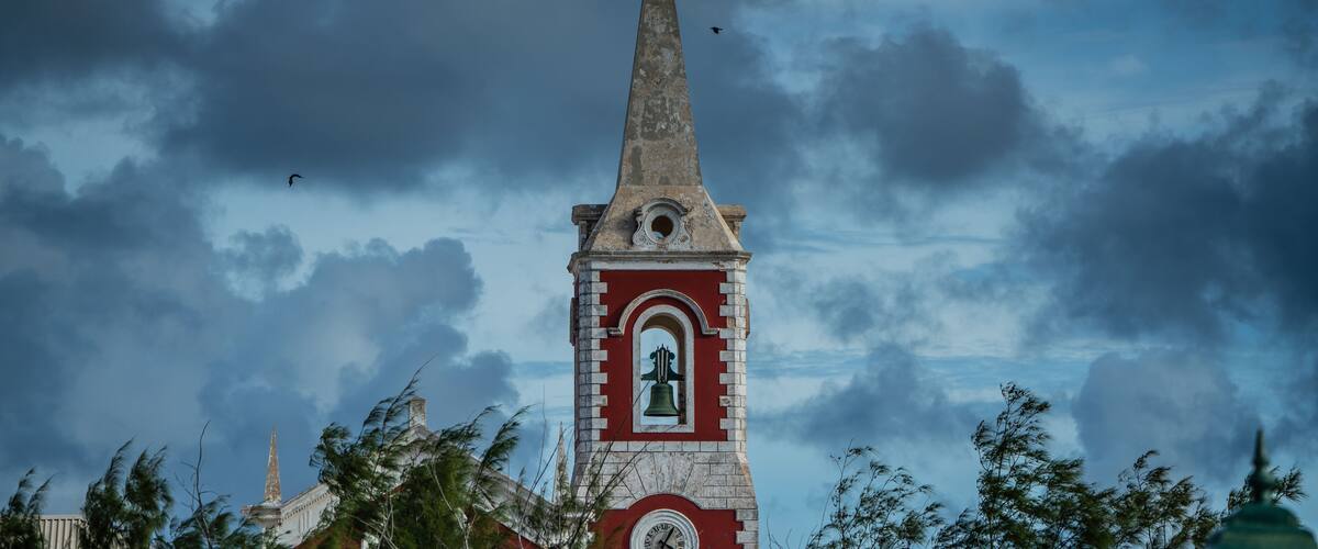 A church tower with a clock and a bell, surrounded by trees and a cloudy sky.