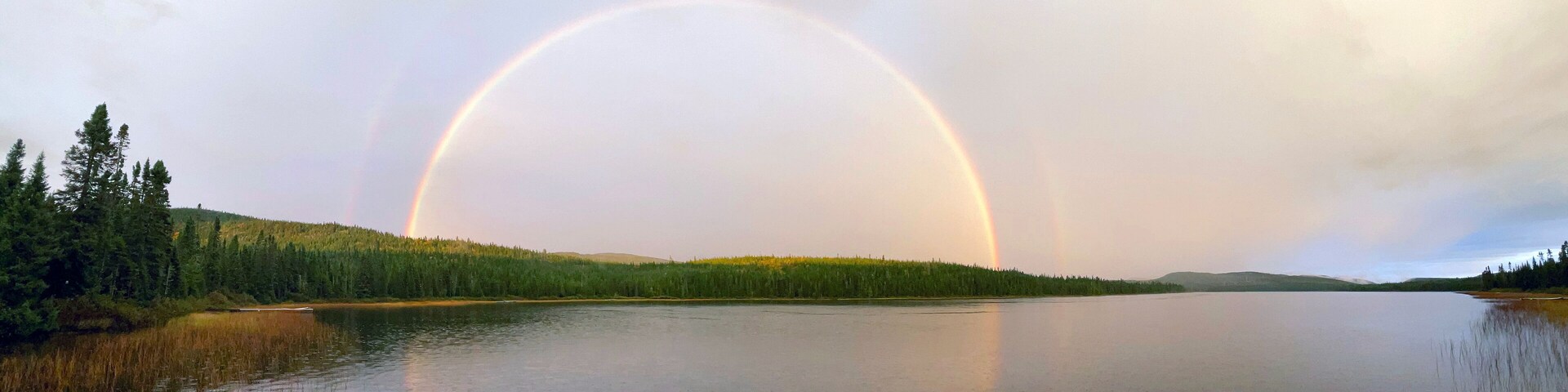 Um lago com um arco-íris sobre ele, rodeado por uma floresta densa e colinas.