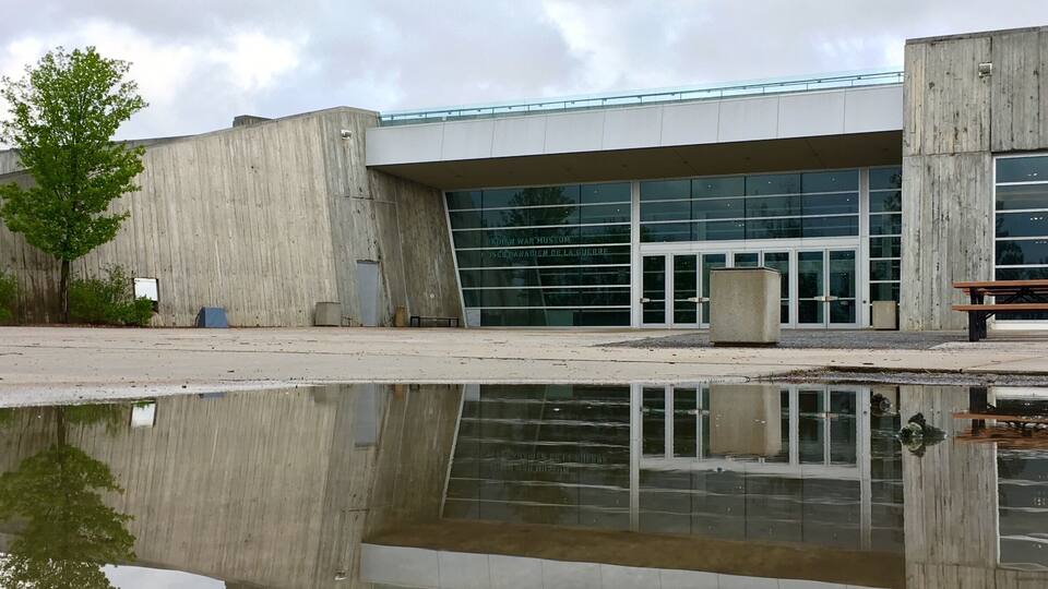 A modern building with a glass facade and a concrete exterior. There is a reflection of the building in a puddle on the ground.