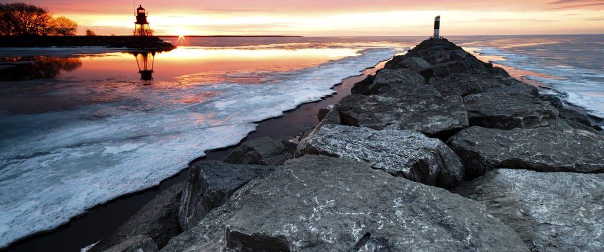 A rocky breakwater at sunset with a lighthouse and a solitary figure on a rock.