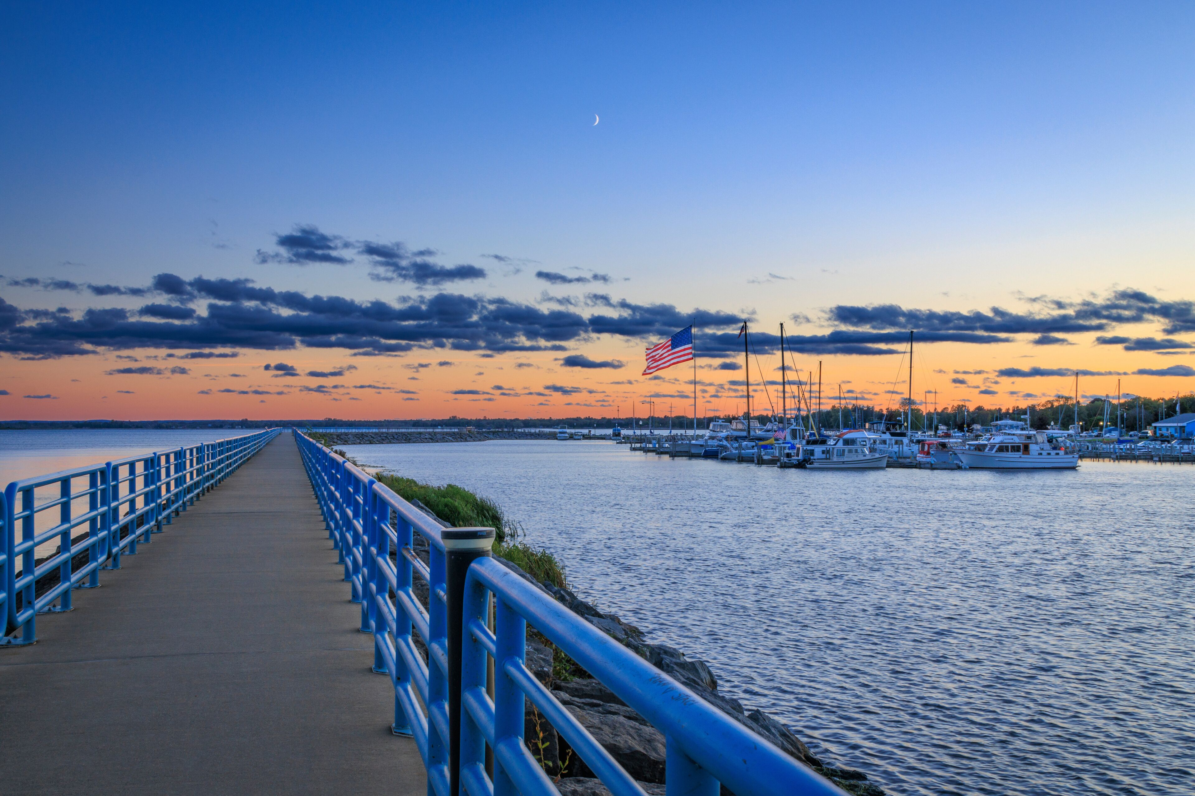 A waterfront promenade with a blue railing, a marina with boats, and a sunset sky.
