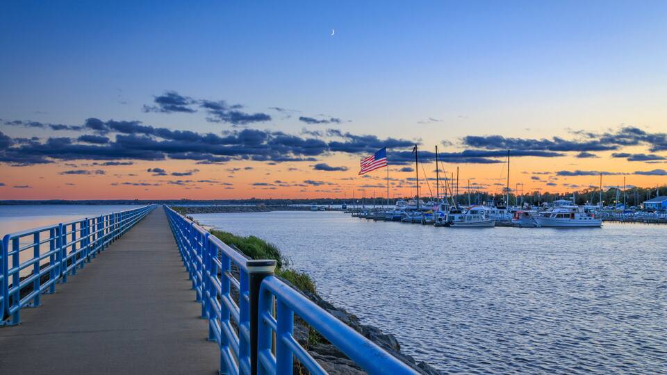 A waterfront promenade with a blue railing, a marina with boats, and a sunset sky.