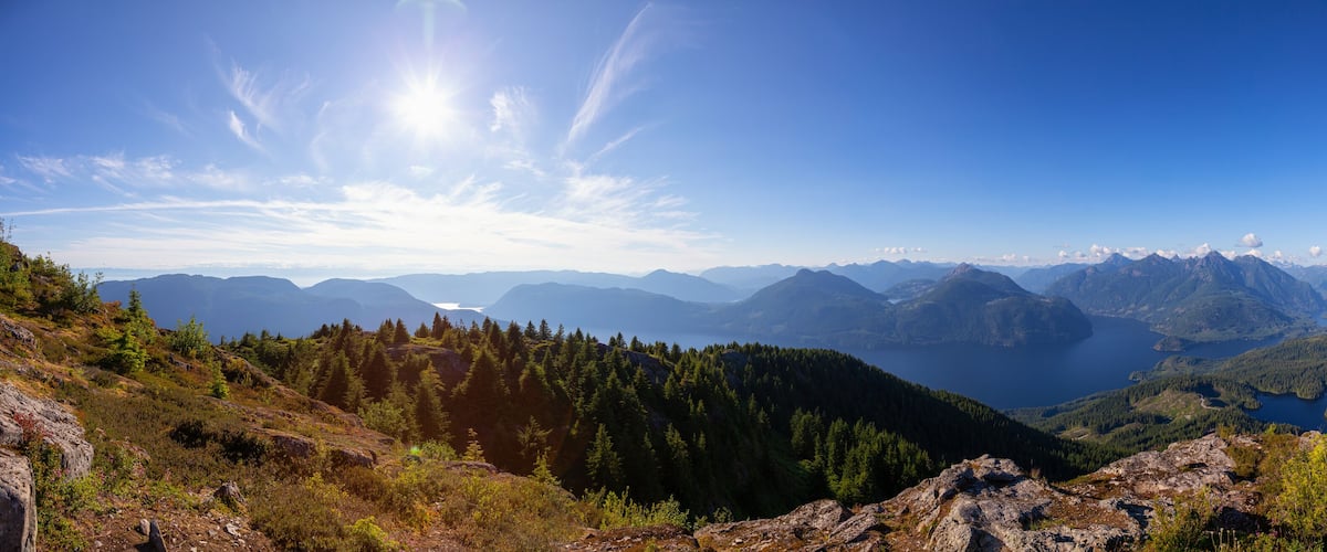 A mountainous landscape with dense forests, rocky terrain, and a lake in the distance.