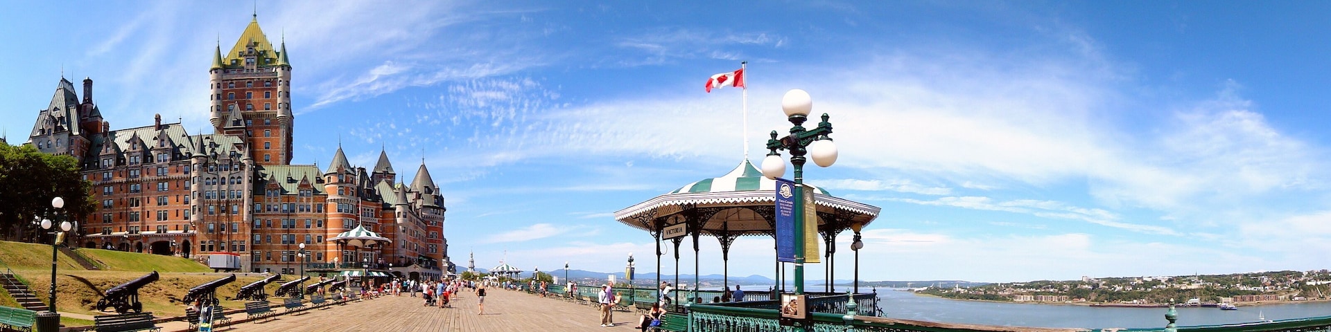 Um edifício histórico com uma torre, um calçadão à beira-mar e um coreto com a bandeira do Canadá.