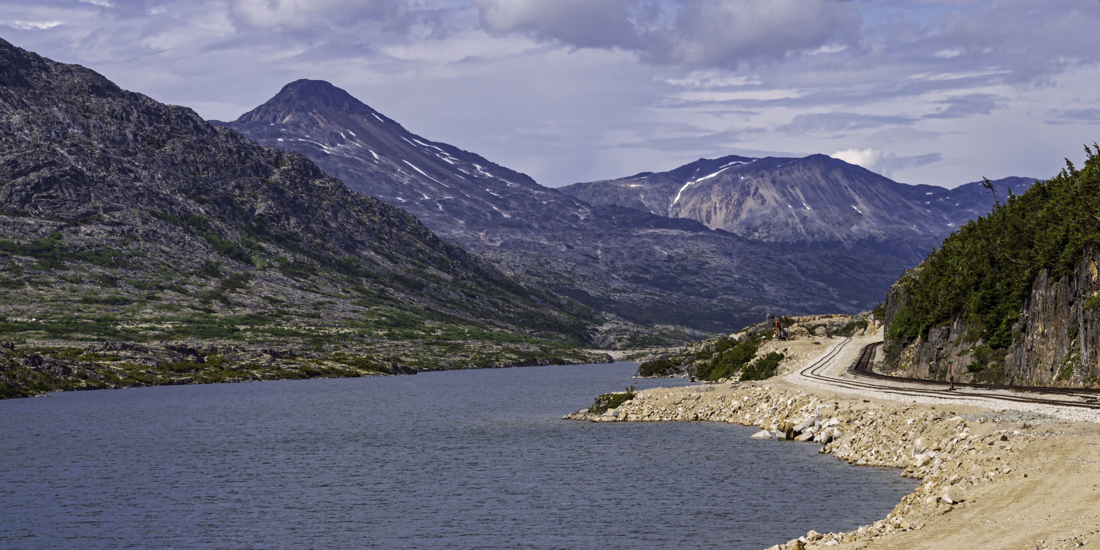 A mountainous landscape with a lake, a railway track, and a rocky shoreline.