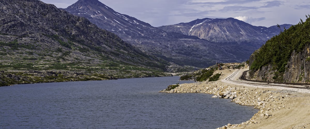A mountainous landscape with a lake, a railway track, and a rocky shoreline.