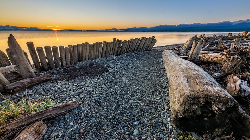 A pebbled beach with driftwood and a wooden fence, overlooking a calm lake with mountains in the distance.