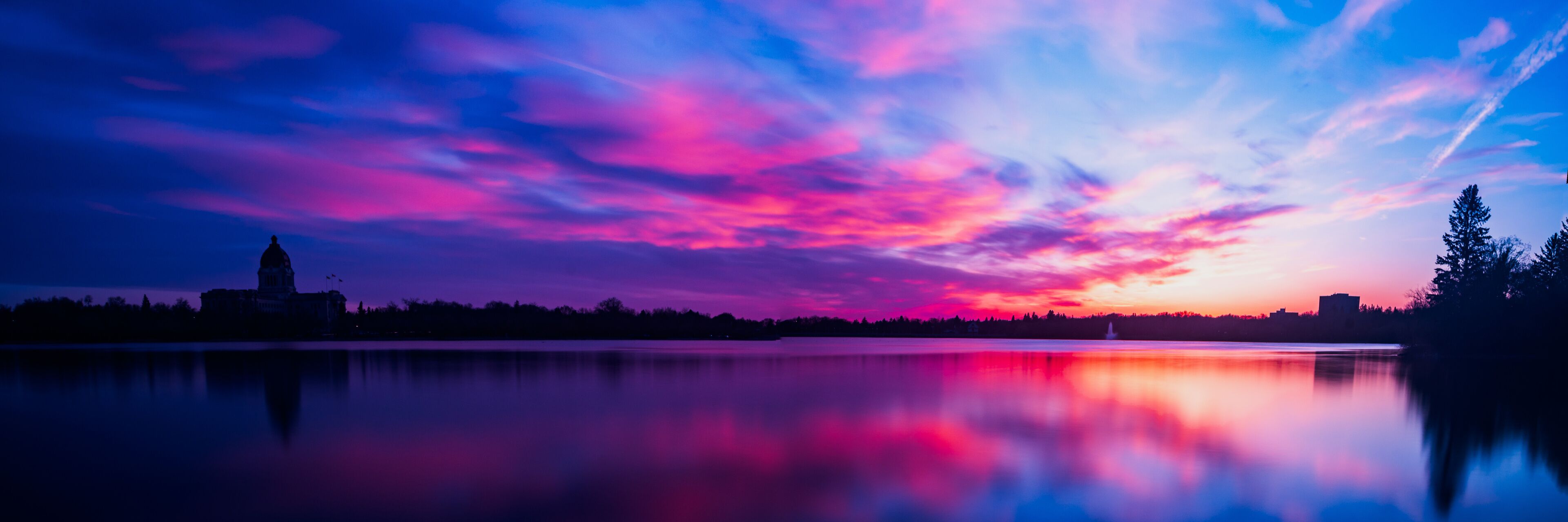 A sunset over a calm lake with vibrant pink and purple clouds, a silhouette of a city skyline, and a distant building.