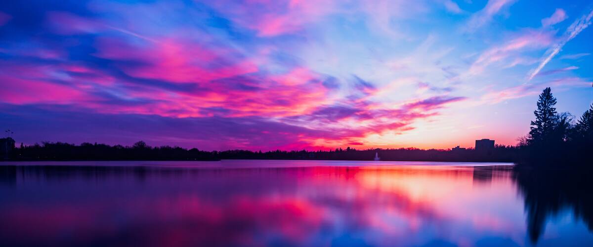 A sunset over a calm lake with vibrant pink and purple clouds, a silhouette of a city skyline, and a distant building.