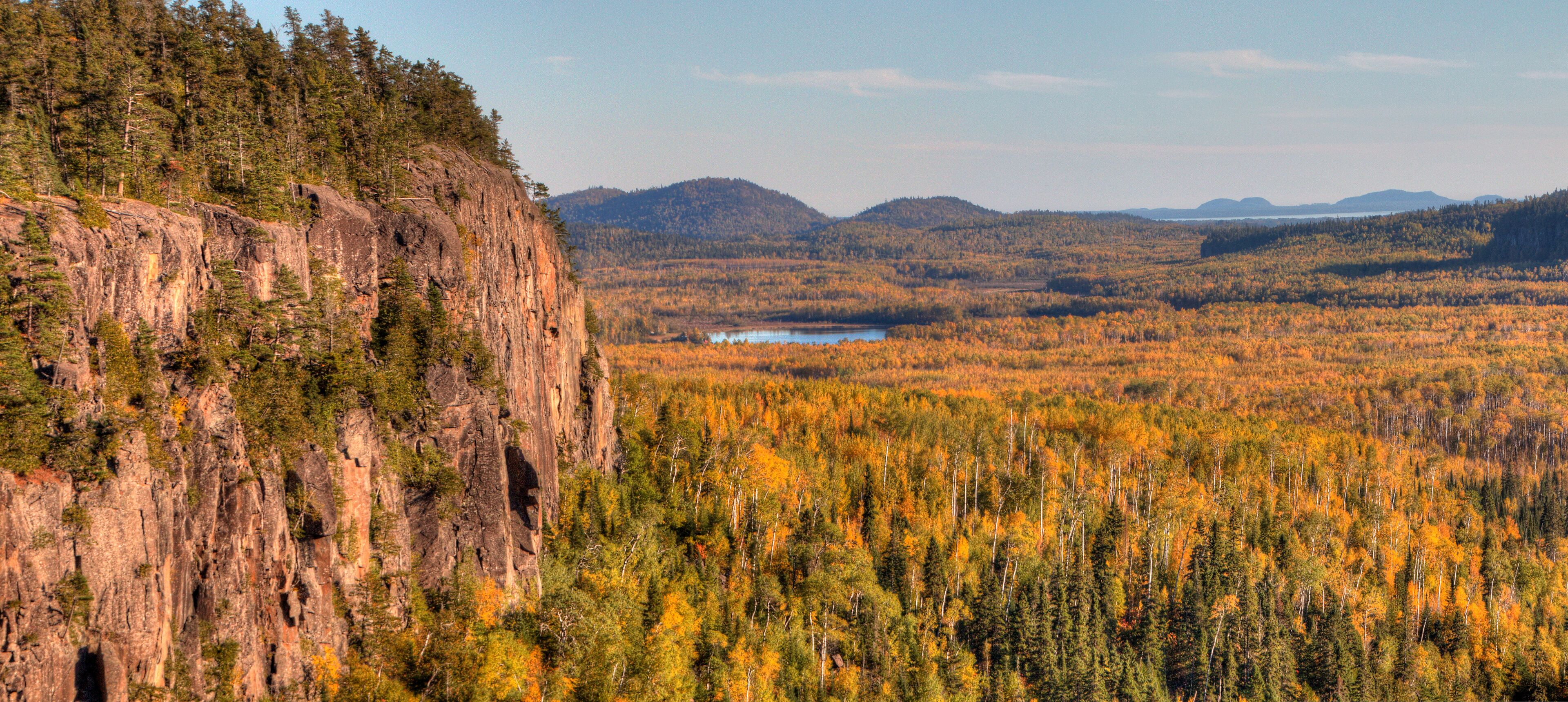 A forested valley with a rocky cliff and a lake in the distance.