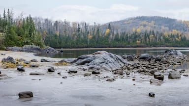 Rocky shoreline by a calm lake with forested hills in the background under a cloudy sky. Thunder Bay, Ontario, Canada
