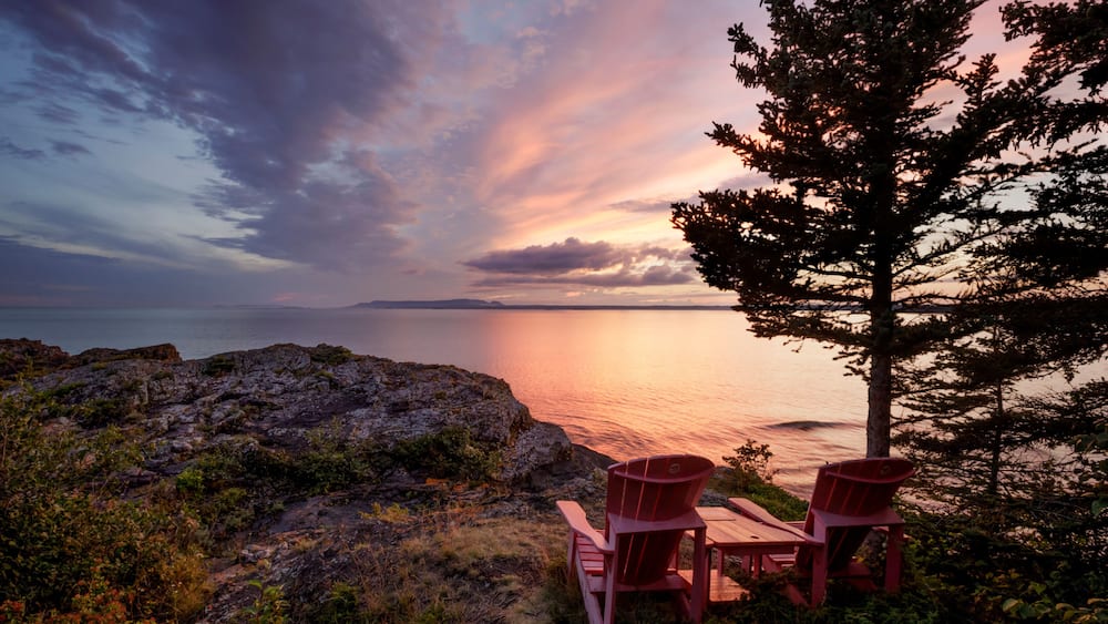 Two red adirondack chairs on a rocky ridge with a view of the Sleeping Giant from Porphryry Island; Porphryry Island, Ontario, Canada