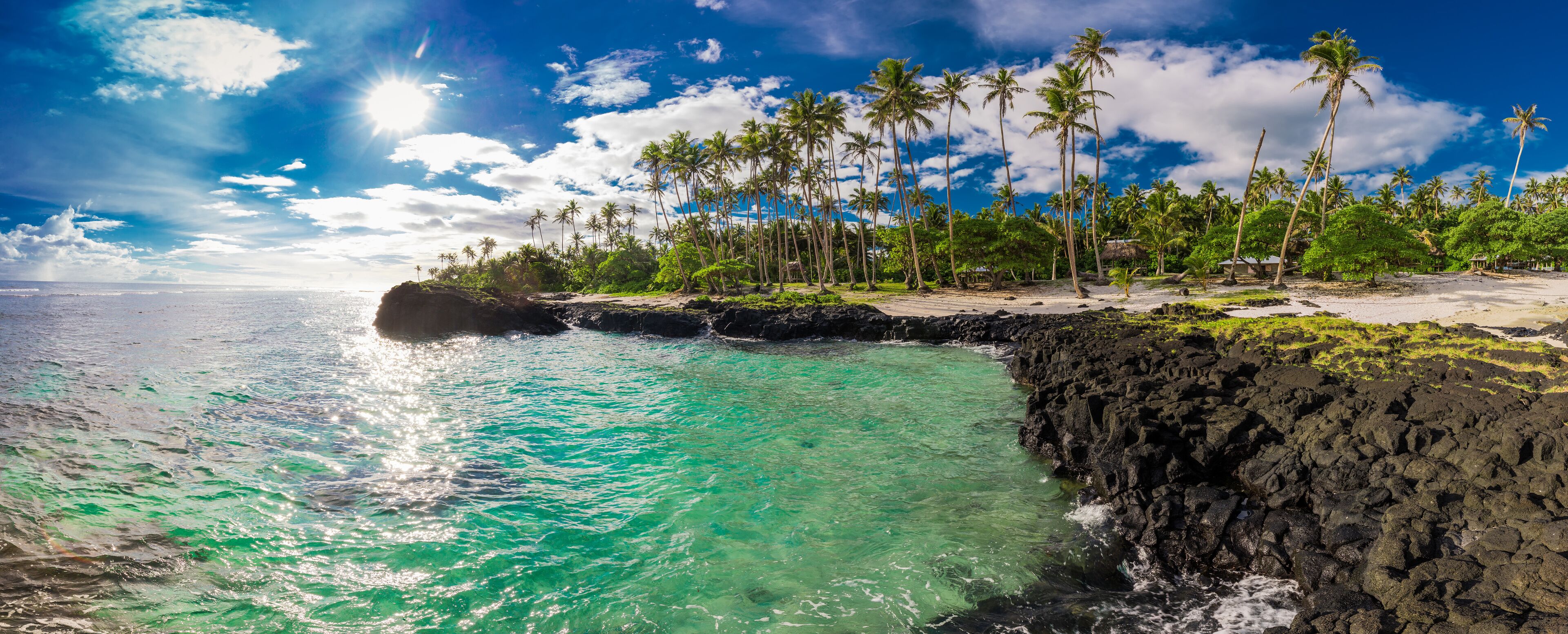 A tropical beach with clear turquoise water, palm trees, and rocky cliffs.
