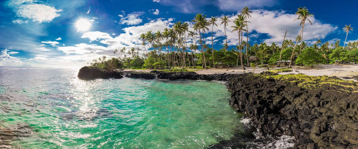 A tropical beach with clear turquoise water, palm trees, and rocky cliffs.