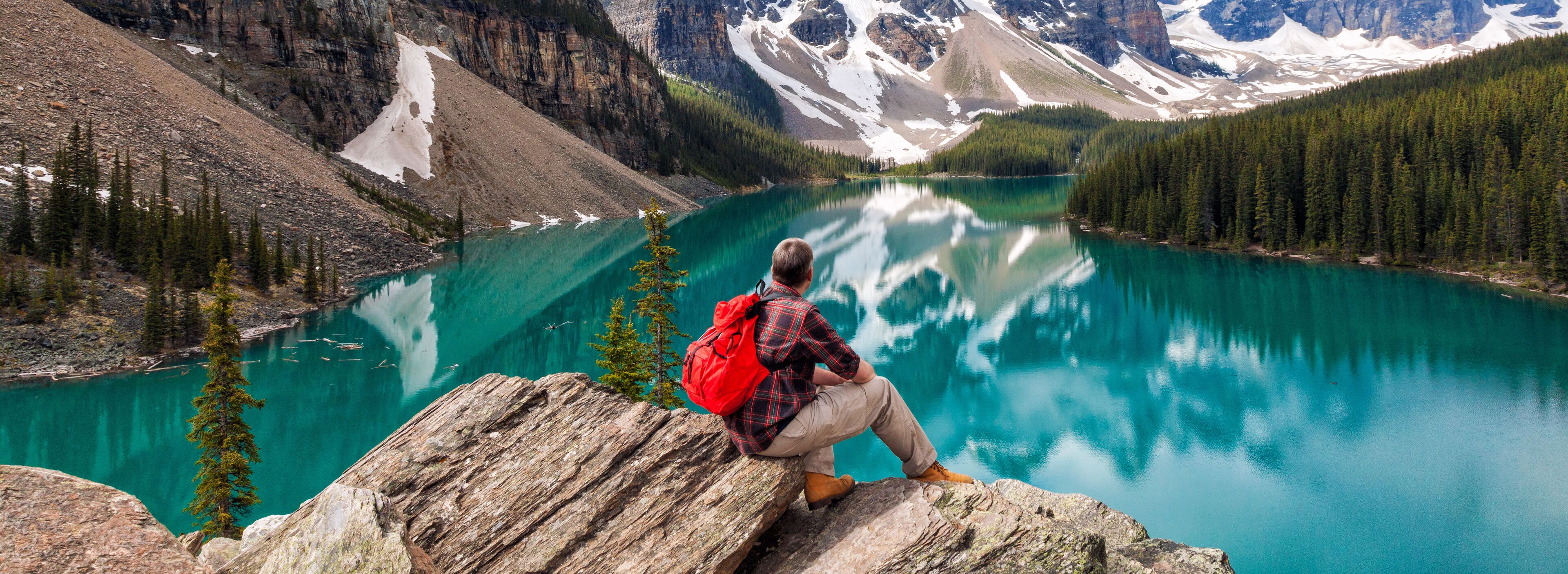 A person sitting on a rock overlooking a turquoise lake surrounded by mountains and dense forests.