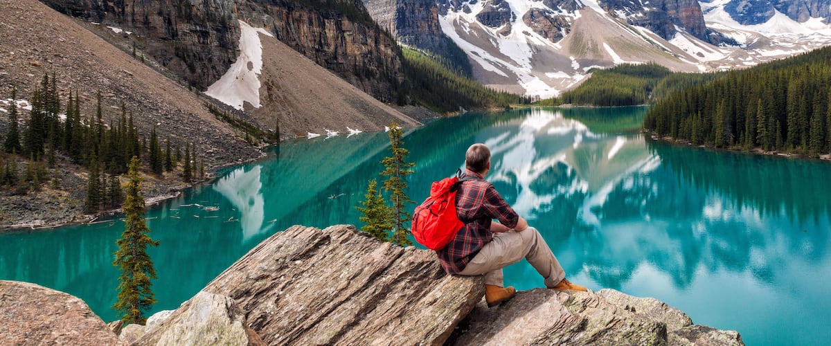 A person sitting on a rock overlooking a turquoise lake surrounded by mountains and dense forests.