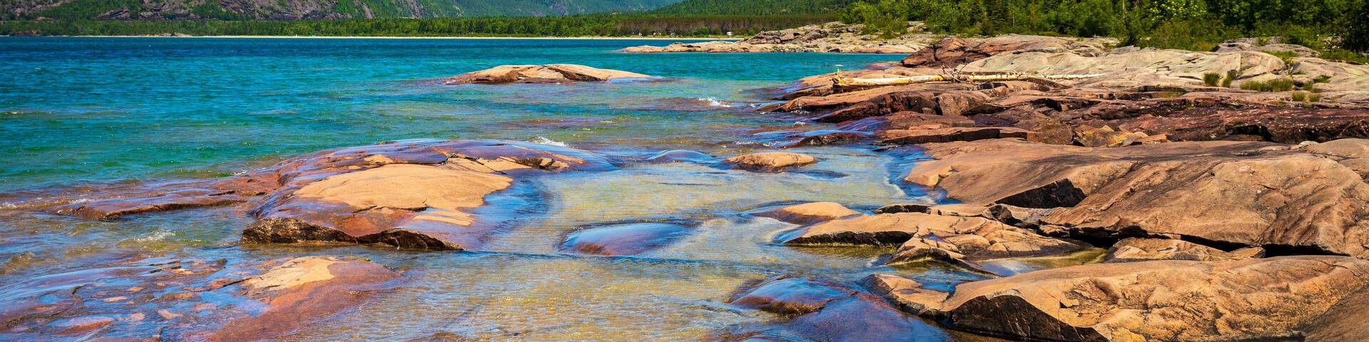 A rocky shoreline with clear turquoise water, surrounded by green hills and dense forest.