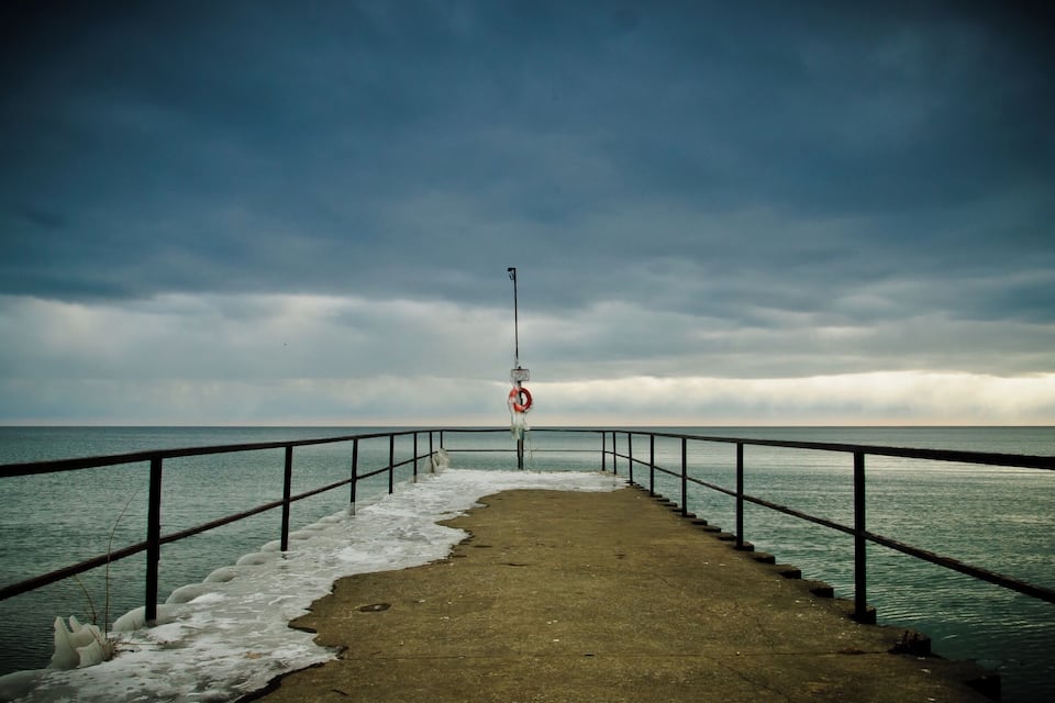 Un muelle de hormigón con barandilla metálica que se adentra en un mar tranquilo bajo un cielo nublado.