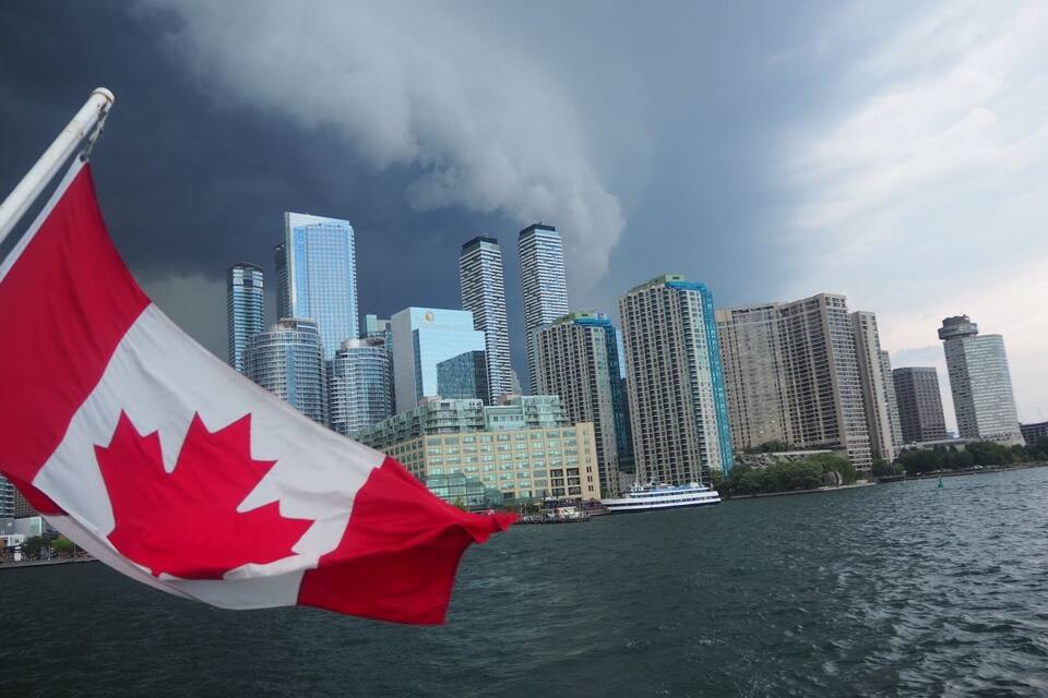 A Canadian flag waving in front of a city skyline with tall buildings and a body of water.