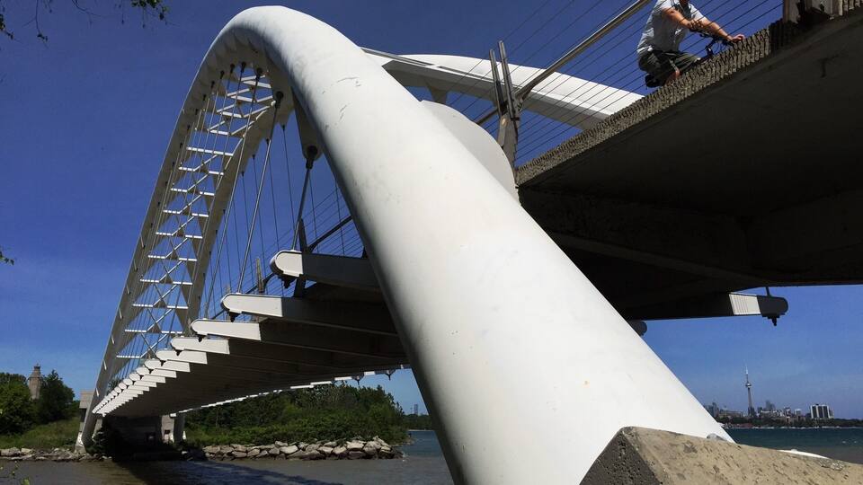 Un puente moderno con un diseño curvo, una persona en la pasarela y un horizonte urbano de fondo.