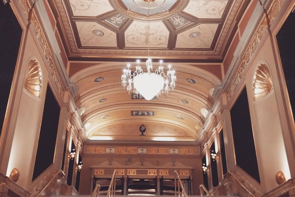 Uma escadaria imponente leva a um teto ricamente decorado de um teatro, com um candelabro.