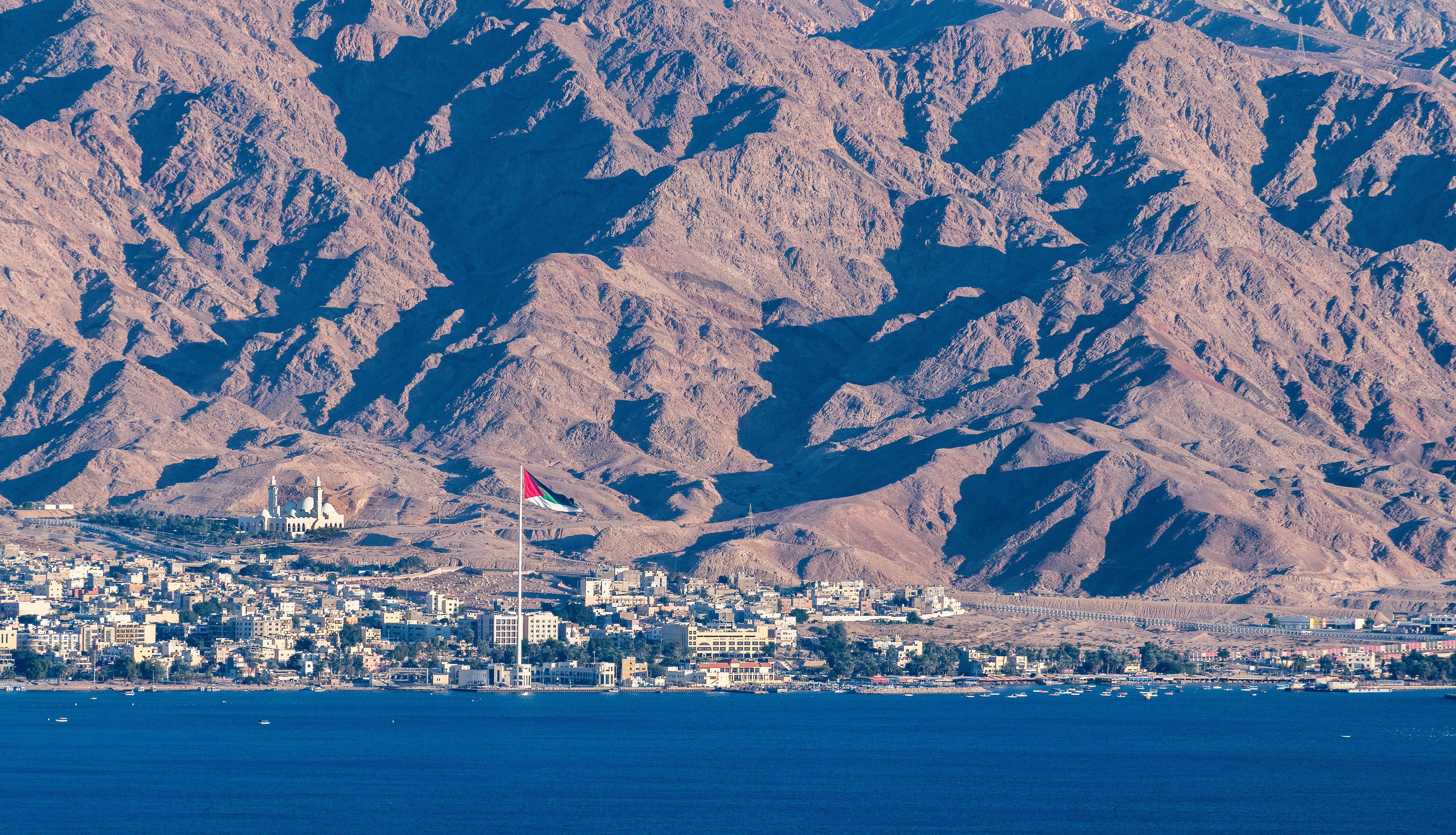 A coastal city with a prominent mosque and flag, surrounded by rugged mountains.