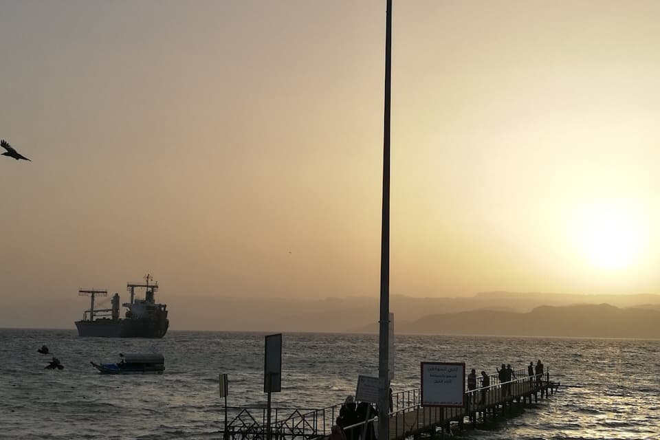 Een zonsondergang boven een kalme zee, met een steiger, mensen die aan de kust zitten en een schip in de verte.