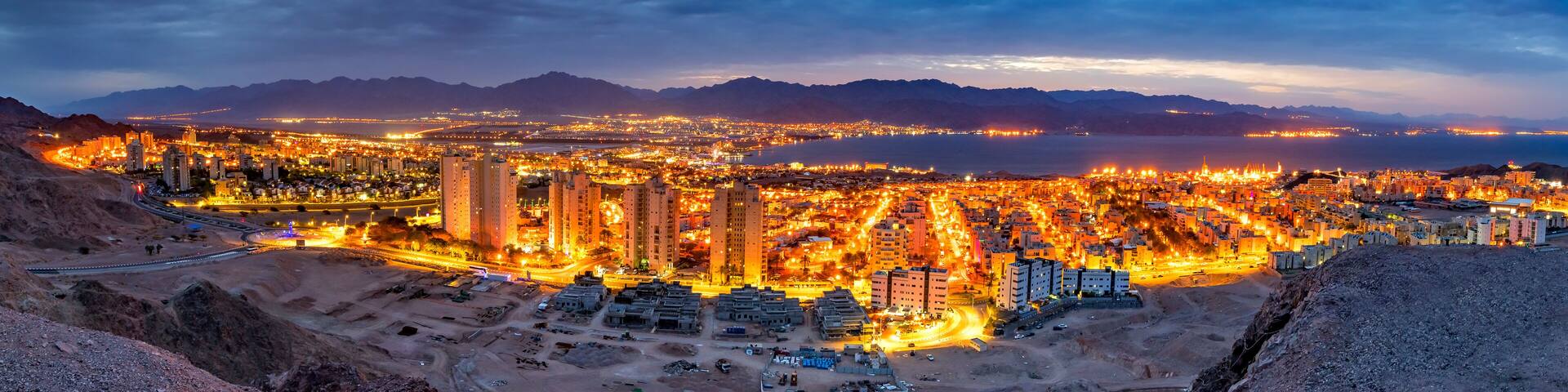 A cityscape at dusk with illuminated buildings and a mountainous backdrop.