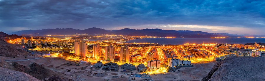 A cityscape at dusk with illuminated buildings and a mountainous backdrop.